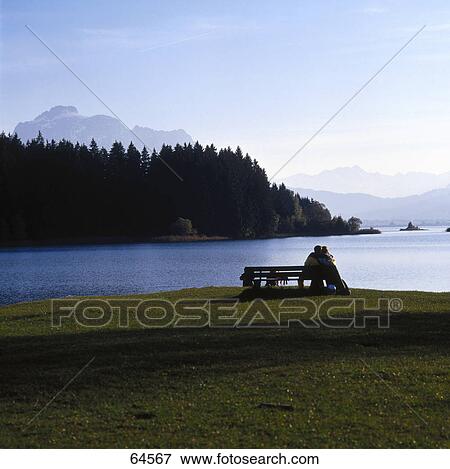 Stock Photo - Rear view of couple sitting on bench at lakeside, Bavaria, Germany . Fotosearch