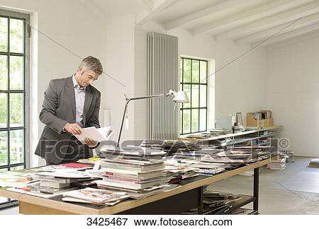 Stock Photo - Man standing in front of a table with magazines and books and reading a book . Fotosearch