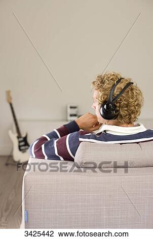 Stock Image - Man with headphones sitting on a couch, e-guitar in the background, rear view . Fotosearch