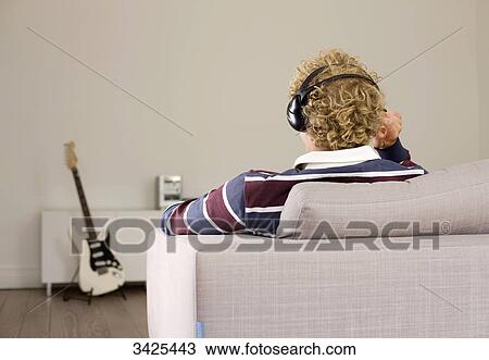 Stock Image - Man with headphones sitting on a couch, e-guitar in the background, rear view . Fotosearch