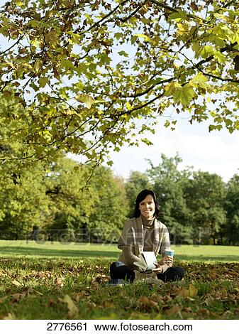 Stock Image - Portrait of mature woman holding book and smiling in park . Fotosearch