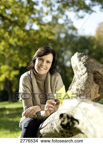 Picture - Portrait of mature woman sitting on fallen tree and smiling . Fotosearch