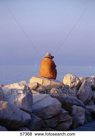 Stock Photo - Rear view of a person sitting on rocks at the coast, Piran, Slovenia . Fotosearch