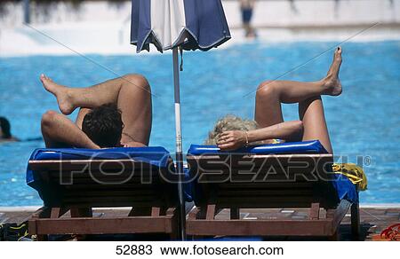 Stock Image - Rear view of couple resting on lounge chairs at poolside . Fotosearch