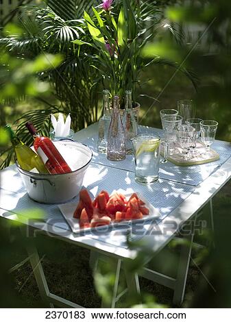Stock Image - Watermelon slices with beverages and potted plant on table in garden . Fotosearch
