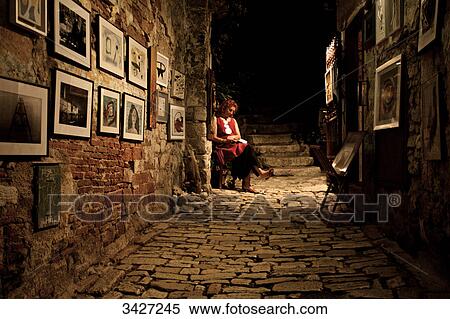 Stock Photography - Woman sitting in an alley, pictures hanging on the wall, Croatia . Fotosearch
