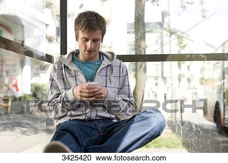 Stock Image - Young man sitting in a bus stop and using a mp3-player, low angle view . Fotosearch
