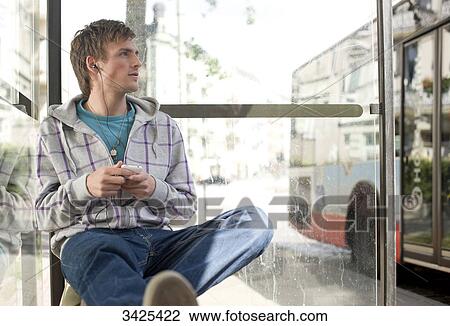 Stock Image - Young man sitting in a bus stop and using a mp3-player, low angle view . Fotosearch