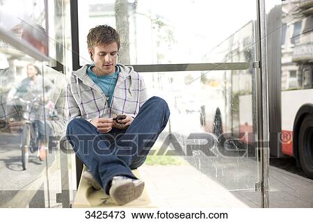 Stock Image - Young man sitting in a bus stop and using a mp3-player, low angle view . Fotosearch