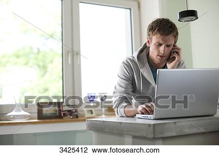 Stock Image - Young man standing at a counter and using mobile phone and laptop, low angle view . Fotosearch