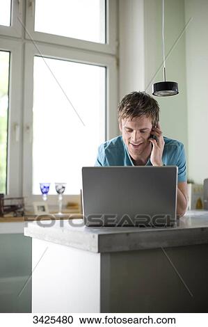 Young man standing at a counter and using mobile phone and laptop, low angle view View Large Photo Image Stock Image - Young man standing at a counter and using mobile phone and laptop, low angle view . Fotosearch