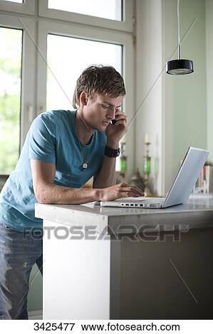 Young man standing at a counter and using mobile phone and laptop, low angle view View Large Photo Image Stock Photo - Young man standing at a counter and using mobile phone and laptop, low angle view . Fotosearch