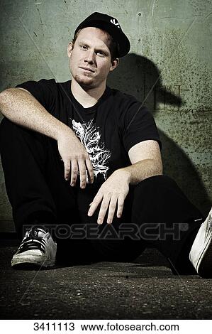 Stock Image - Young man with baseball cap sitting in front of a gate, portrait . Fotosearch