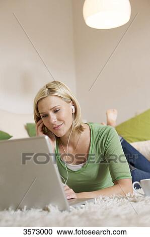 Stock Image - Young woman lying on rug with cup of coffee, earbuds and laptop . Fotosearch