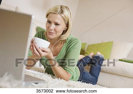 Stock Image - Young woman lying on rug with cup of coffee, earbuds and laptop . Fotosearch