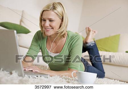 Stock Photo - Young woman lying on rug with cup of coffee, earbuds and laptop . Fotosearch