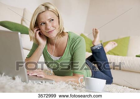 Stock Photo - Young woman lying on rug with cup of coffee, earbuds and laptop . Fotosearch