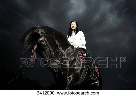 Stock Image - Young woman riding on a black horse . Fotosearch