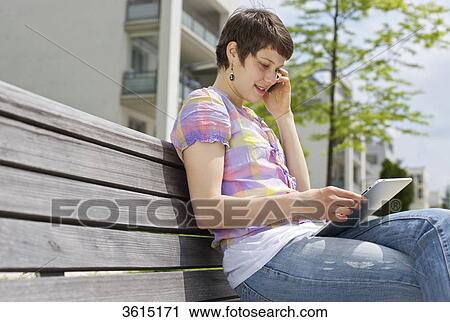 Stock Image - Young woman with cell phone and iPad on a bench . Fotosearch