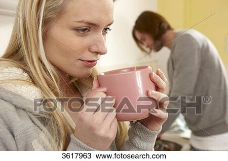 Stock Image - Young woman drinking coffee in kitchen with man in background . Fotosearch