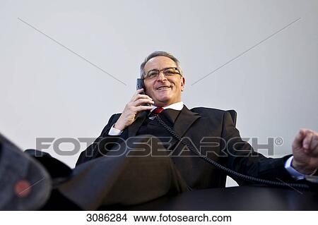 Businessman talking on phone with feet up on table, low angle view View Large Photo Image Picture - Businessman talking on phone with feet up on table, low angle view . Fotosearch