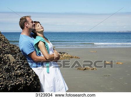 Picture - Couple embracing on the beach, leaning against a rock, side view . Fotosearch