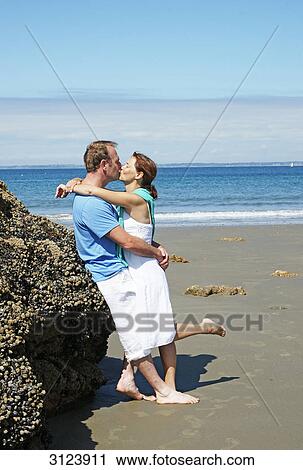 Stock Image - Couple kissing on the beach, leaning against a rock, side view . Fotosearch