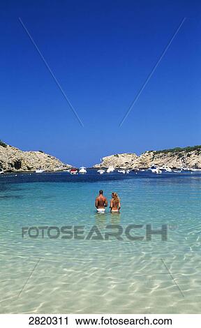 Stock Image - Rear view of couple standing in shallow water, Cala Moli, Ibiza, Balearic Islands, Spain . Fotosearch