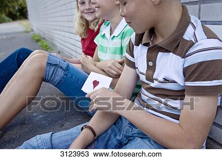 Stock Image - Three children sitting on the ground, boy holding a card with a heart on it . Fotosearch
