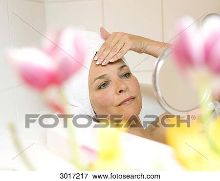 Stock Photo - Woman lying in a bath tub and looking at herself in a hand mirror, close-up . Fotosearch