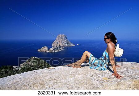 Stock Image - Woman sitting on cliff looking at sea, Ibiza, Balearic Islands, Spain . Fotosearch