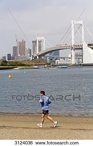 Stock Photograph - Jogger at the riverbank, Rainbow Bridge and skyline in the background, Tokyo, Japan . Fotosearch