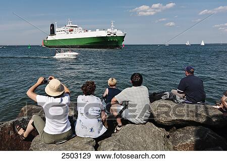 Stock Photo - Tourists sitting on rocks at coast looking at container ship, Warnemunde, Mecklenburg-Vorpommern, Berlin, Germany . Fotosearch