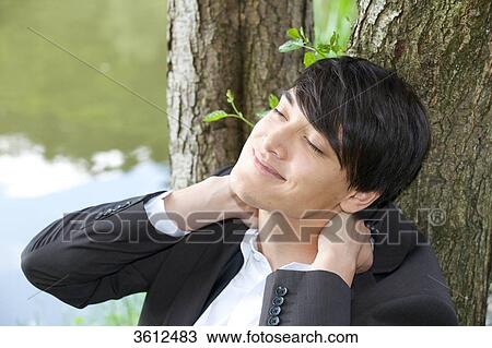 Stock Image - Young businessman leaning against tree trunk . Fotosearch