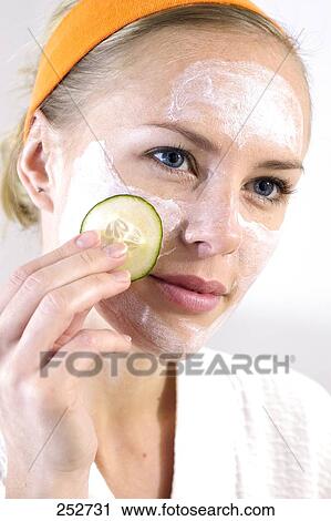 Stock Image - Close-up of young woman applying face mask moisturizer with cucumber . Fotosearch