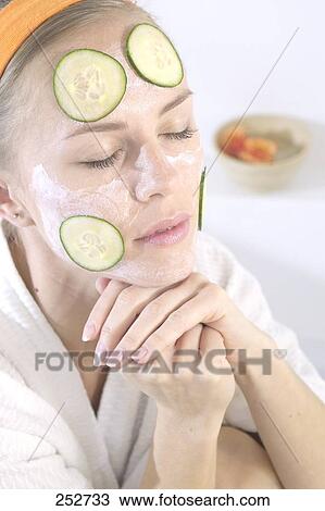 Stock Image - Close-up of young woman with facial mask and cucumber slices on her face . Fotosearch