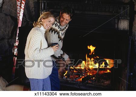 Couple In Ski Lodge Standing By Fireplace Man Stoking Fire
