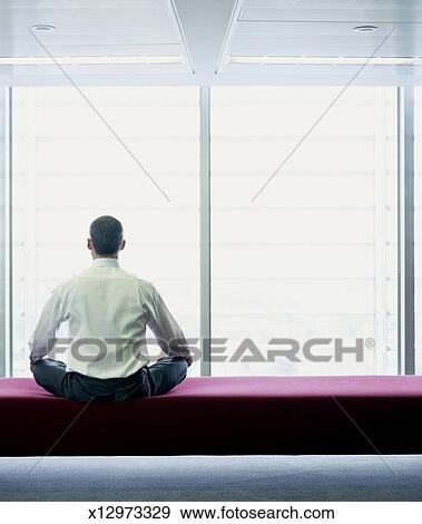 Business Man Sitting Cross Legged On Bench By Window Rear View