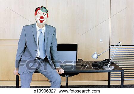 Businessman Sitting At Edge Of Desk Wearing Clown Mask Stock Photo