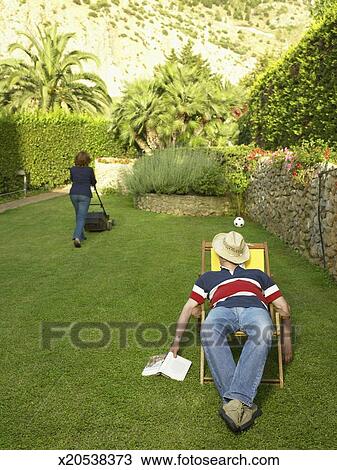 Mature man relaxing in deckchair in garden, mature woman mowing lawn