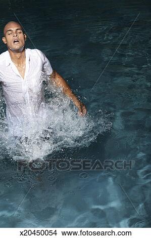 Young man in wet shirt in swimming pool, jumping out of water, elevated