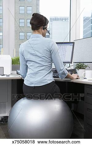 Female Office Worker Sitting On Large Silver Exercise Ball At Desk