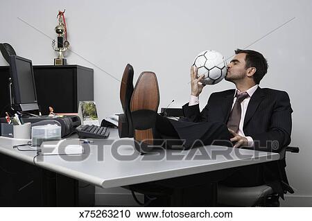 Businessman Sitting With Feet Up On Desk Kissing Soccer Ball