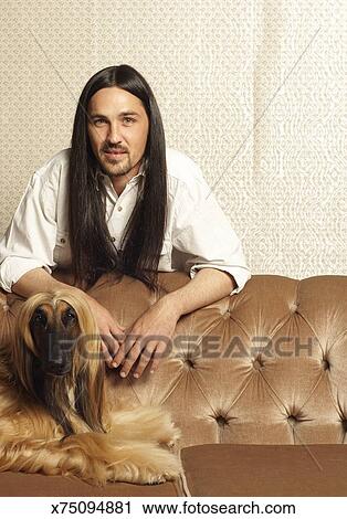 Man With Long Dark Hair Standing By Afghan Hound Lying On Sofa