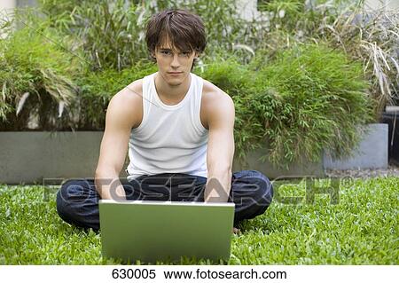 Stock Photography - A young man outdoors sitting on grass using a laptop. Fotosearch
