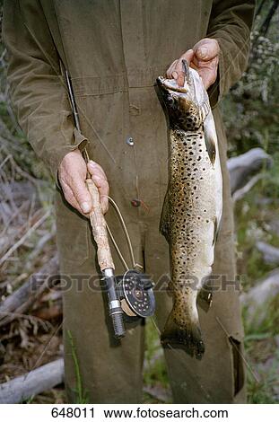 Stock Image - Midsection of a man holding a fishing pole and a fish. Fotosearch