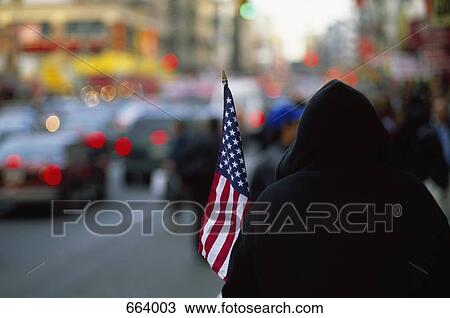 Stock Image - Rear View of a hooded person holding an American flag on a city street. Fotosearch