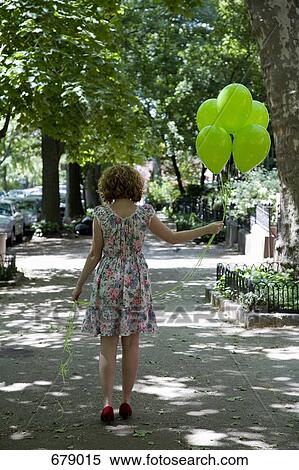 Stock Photography - A young woman walking down a city sidewalk carrying balloons, rear view. Fotosearch