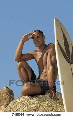 A man sitting on a rock next to a surfboard View Large Photo Image Picture - A man sitting on a rock next to a surfboard. Fotosearch