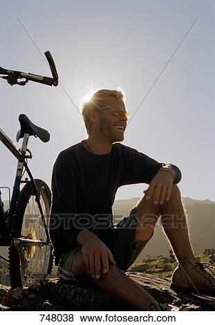 A man sitting on a rock next to a mountain bike View Large Photo Image Stock Photo - A man sitting on a rock next to a mountain bike. Fotosearch